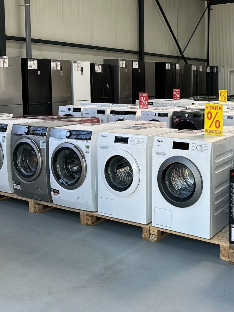 Zu sehen ist eine Reihe von Frontlader-Waschmaschinen mit Energieeffizienz-Kennzeichnungen im Elektromarkt. Im Hintergrund erkennt man noch zahlreiche verschiedene Kühlschränke.