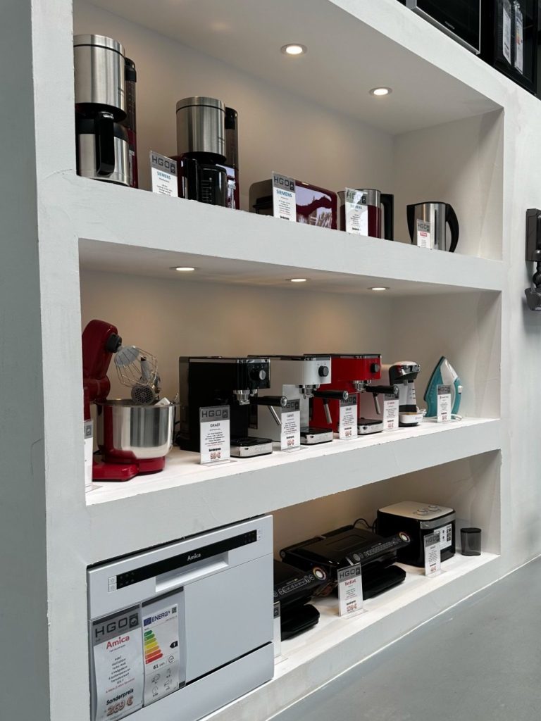 Verschiedene Elektro-Kleingeräte wie Mixer, Küchenmaschinen, Waffeleisen, Toaster und Kaffeemaschinen auf weißen Regalen präsentiert